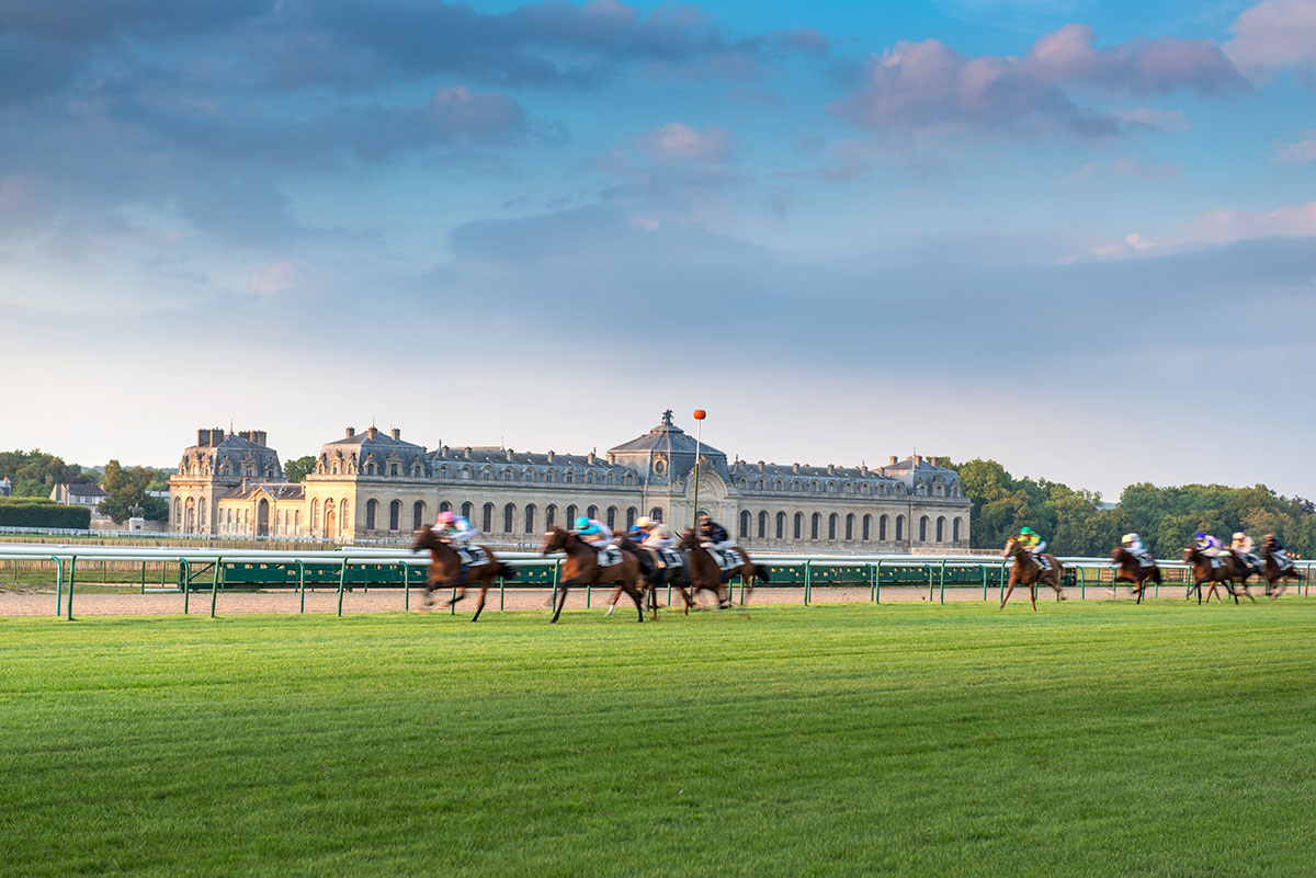Horses racing over green grass in front of a large French chateau.